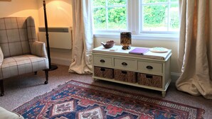 Una habitación con un sillón a cuadros, una lámpara de pie, una ventana con cortinas, una mesita con un cuenco, una vela y un cuaderno.