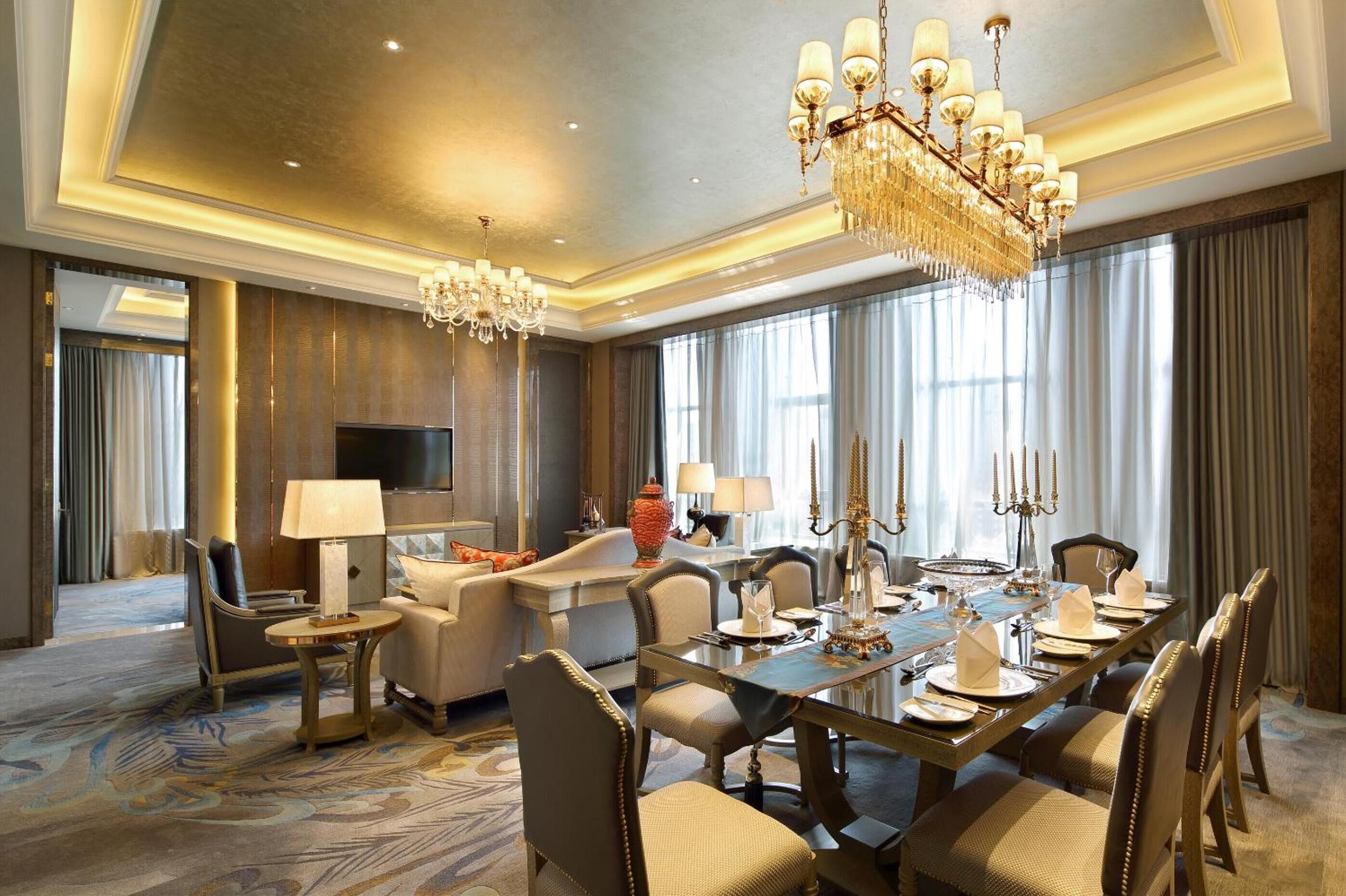 A spacious hotel room with a large dining table set for six, a comfortable seating area with a sofa and armchairs, a flat-screen TV, and a chandelier.