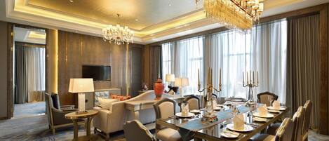 A spacious hotel room with a large dining table set for six, a comfortable seating area with a sofa and armchairs, a flat-screen TV, and a chandelier.