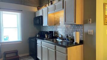 Cocina con estufa, pileta y alacenas. Hay una ventana con persianas y un taburete pequeño.