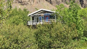 A cabin with a balcony, surrounded by dense greenery and rocky mountains in the background. - Múlakot 5 - Cosy cabin, View the amazing view´s from the porch (Borgarbyggð)