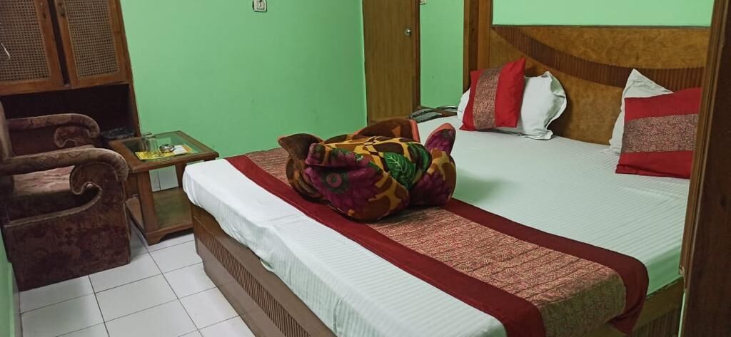 A hotel room with a bed, a wooden wardrobe, a small table, and a colorful bag on the bed.