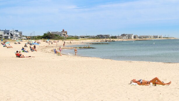 Una spiaggia con persone che prendono il sole e nuotano. Si vedono case e un molo sullo sfondo.