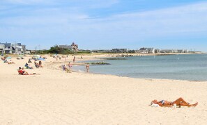 Una playa con gente tomando el sol y nadando. Se ven casas y un muelle al fondo.