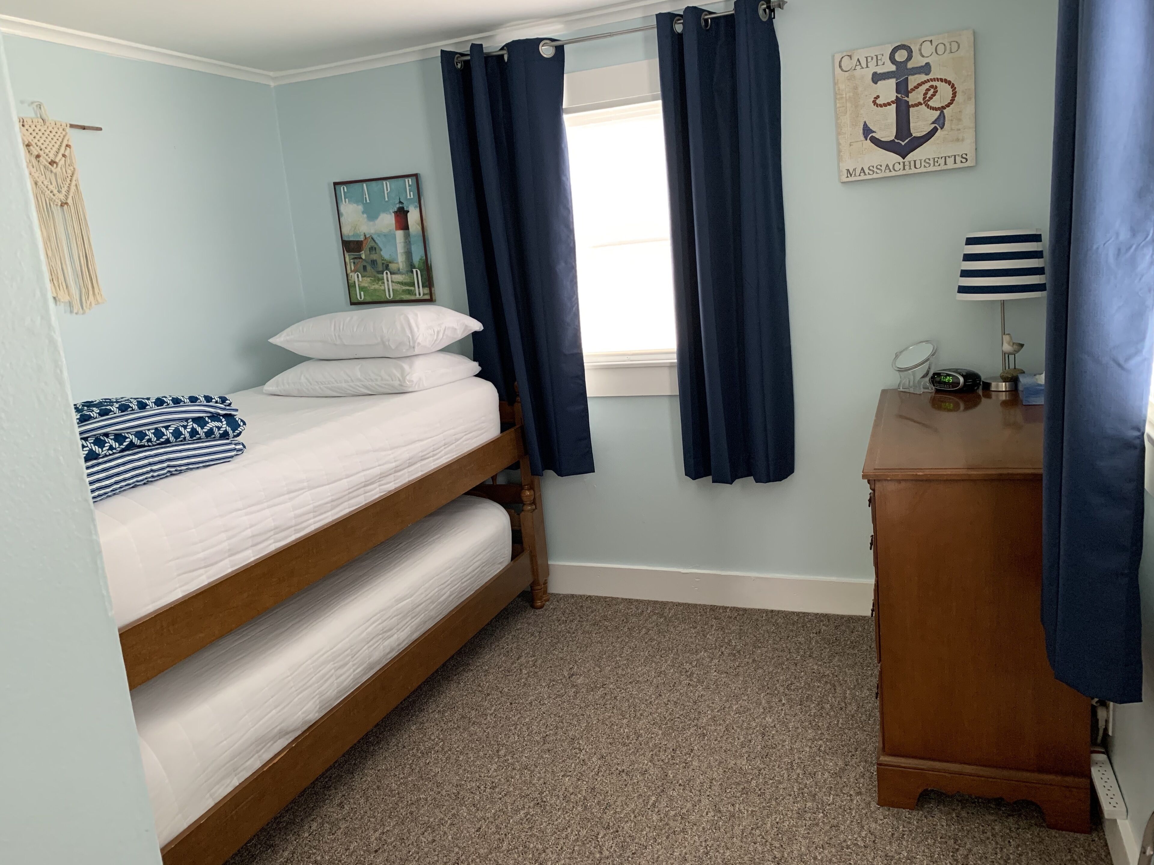 Une chambre avec un lit superposé, une commode en bois, une table de chevet avec une lampe, un tableau encadré et une fenêtre avec des rideaux.