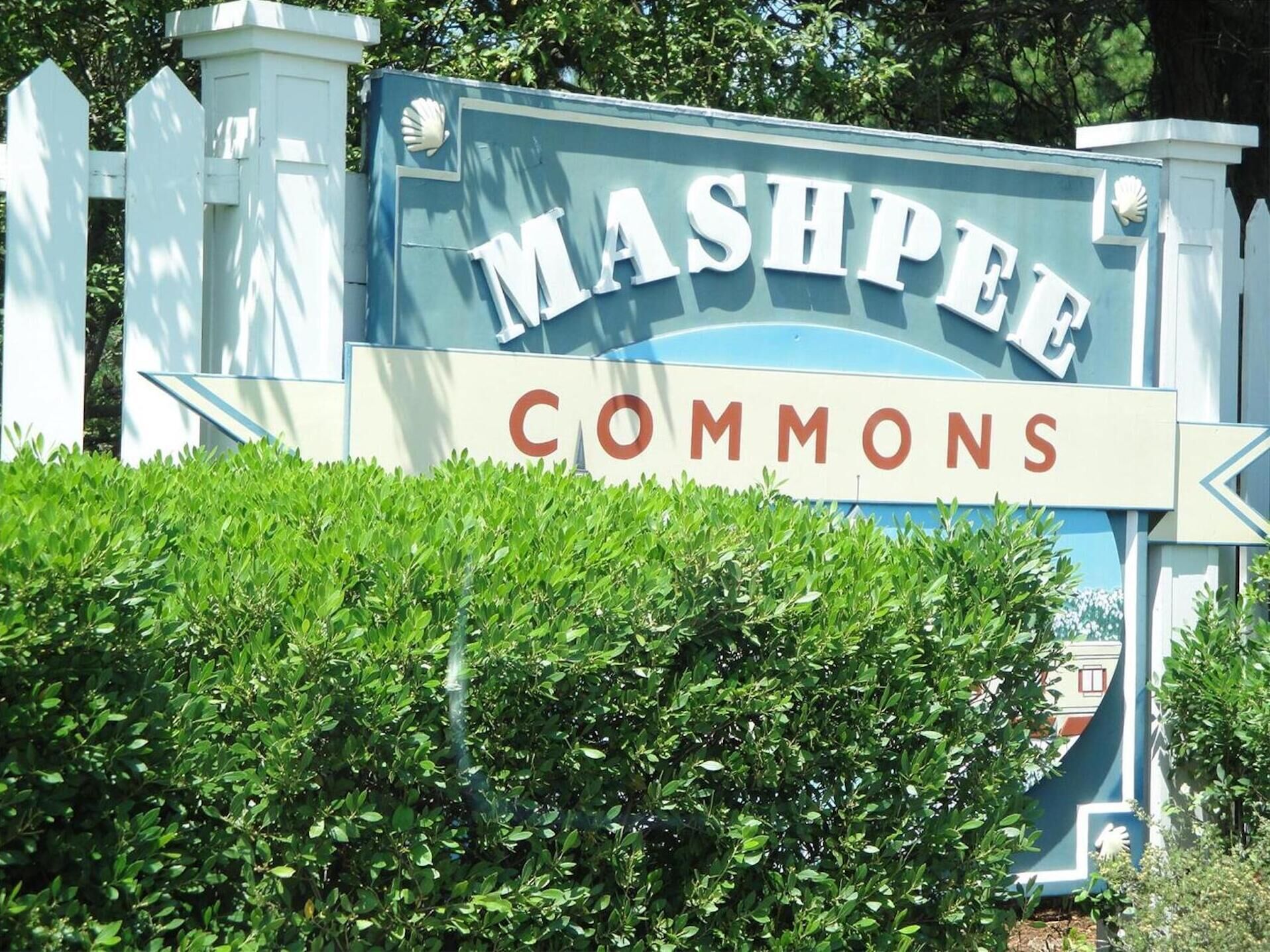 A sign for Mashpee Commons with a white fence and greenery in the foreground.