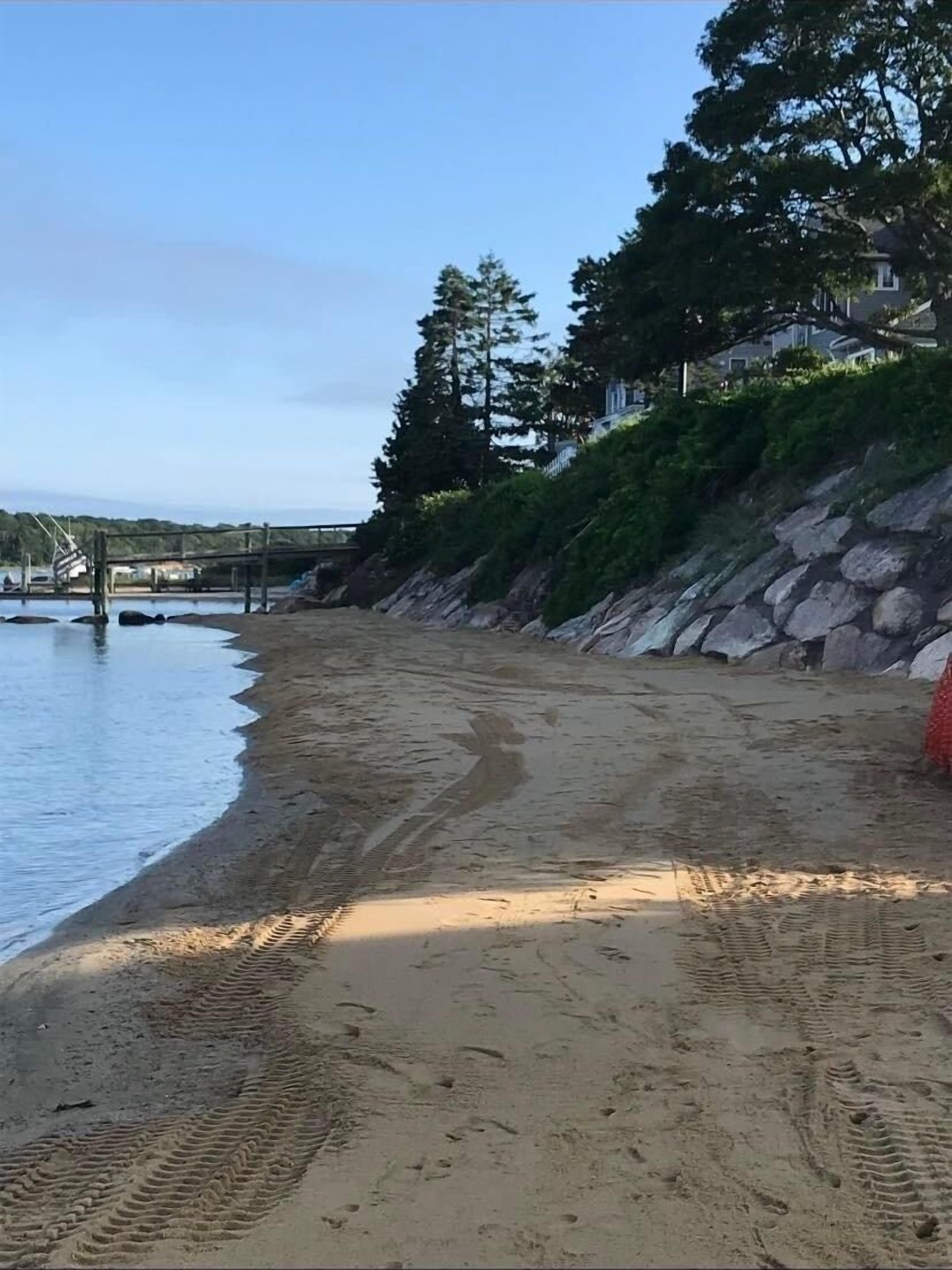 A sandy beach with footprints leading to the water, a rocky hillside, and a bridge in the background.