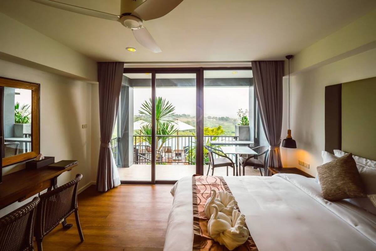 Ein Hotelzimmer mit Bett, Schreibtisch, Stuhl, einem Balkon mit Tisch und Stühlen sowie Blick auf Grünflächen.