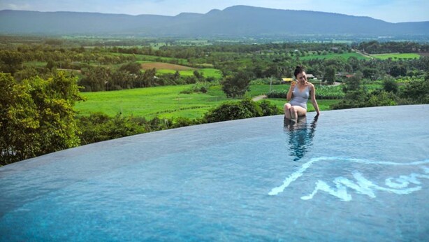 Una persona sentada en el borde de una piscina infinita azul cristalina, con una vista panorámica de campos verdes y montañas al fondo.