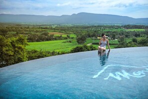A person sitting on the edge of a clear blue infinity pool with a scenic view of green fields and mountains in the background. - Monte Vista Chalet (Pak Chong)