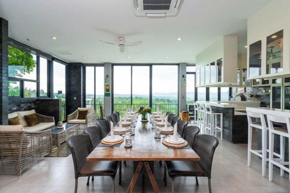 A dining area with a long table set for a meal, surrounded by chairs, with a view of the outdoors.