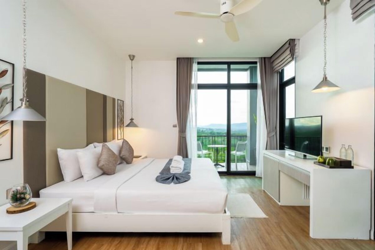 A modern hotel room with a large bed, a desk, a television, and a balcony with a view.