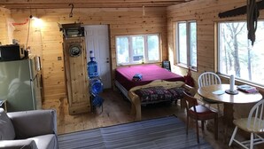 Ein Interieur einer Hütte mit einem Bett, einem Esstisch, Stühlen, einem Kühlschrank, einem Wasserspender und einem Fenster mit Blick auf Bäume.