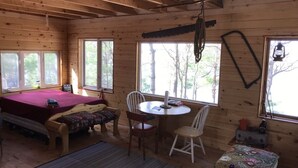 Ein gemütliches Blockhaus-Interieur mit einem Bett, einem Tisch mit Stühlen und großen Fenstern mit Blick auf Bäume.