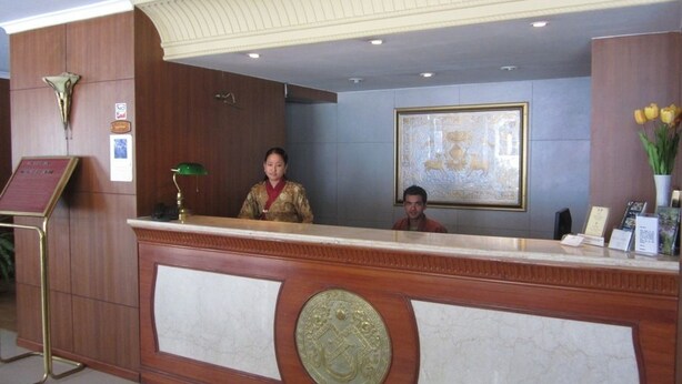 A reception desk with a wooden finish and a marble inlay, a framed artwork on the wall, and a vase with yellow flowers. - Hotel Jumolhari (Thimphu)