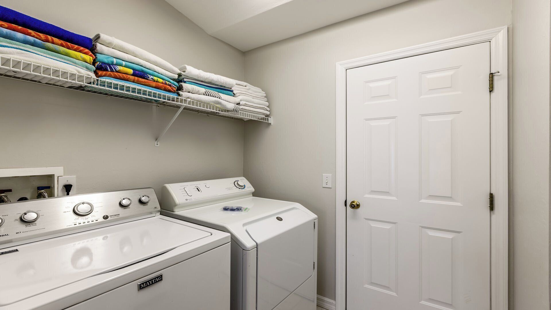 Une buanderie équipée de deux machines à laver et de deux sèche-linge Maytag blancs, d’une porte blanche et d’une étagère au-dessus où sont pliées des serviettes de différentes couleurs.