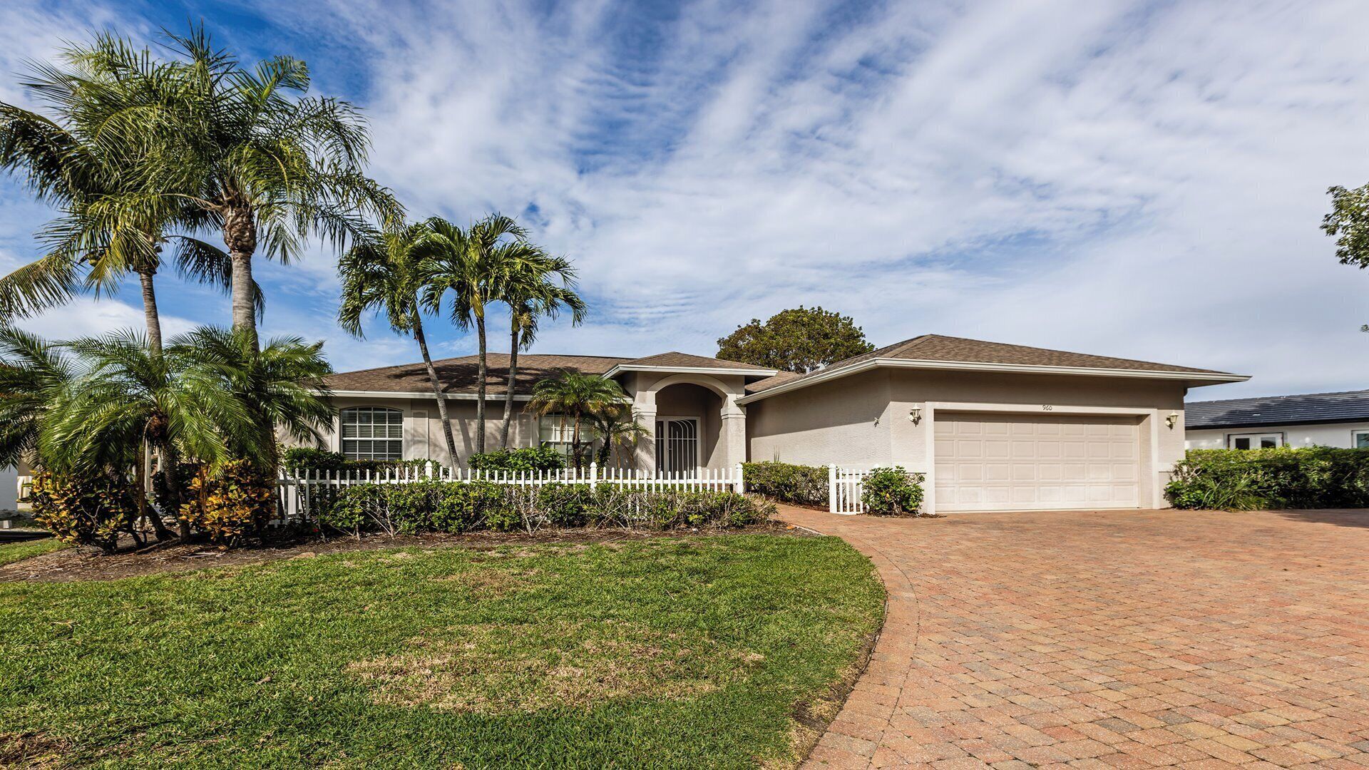 A single-story house with a garage, surrounded by a white picket fence, palm trees, and a well-maintained lawn.