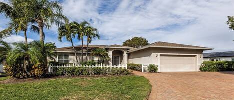 A single-story house with a garage, surrounded by a white picket fence, palm trees, and a well-maintained lawn.