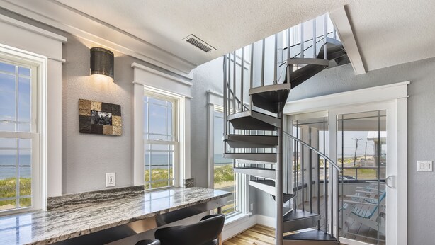 Eine moderne Küche mit Marmorarbeitsplatte, schwarzen Barhocker und einer Wendeltreppe, die zu einem Balkon mit Meerblick führt.