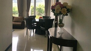 Habitación de hotel con comedor, sofá, mesa con sillas, un jarrón de flores y vistas al exterior con vegetación.