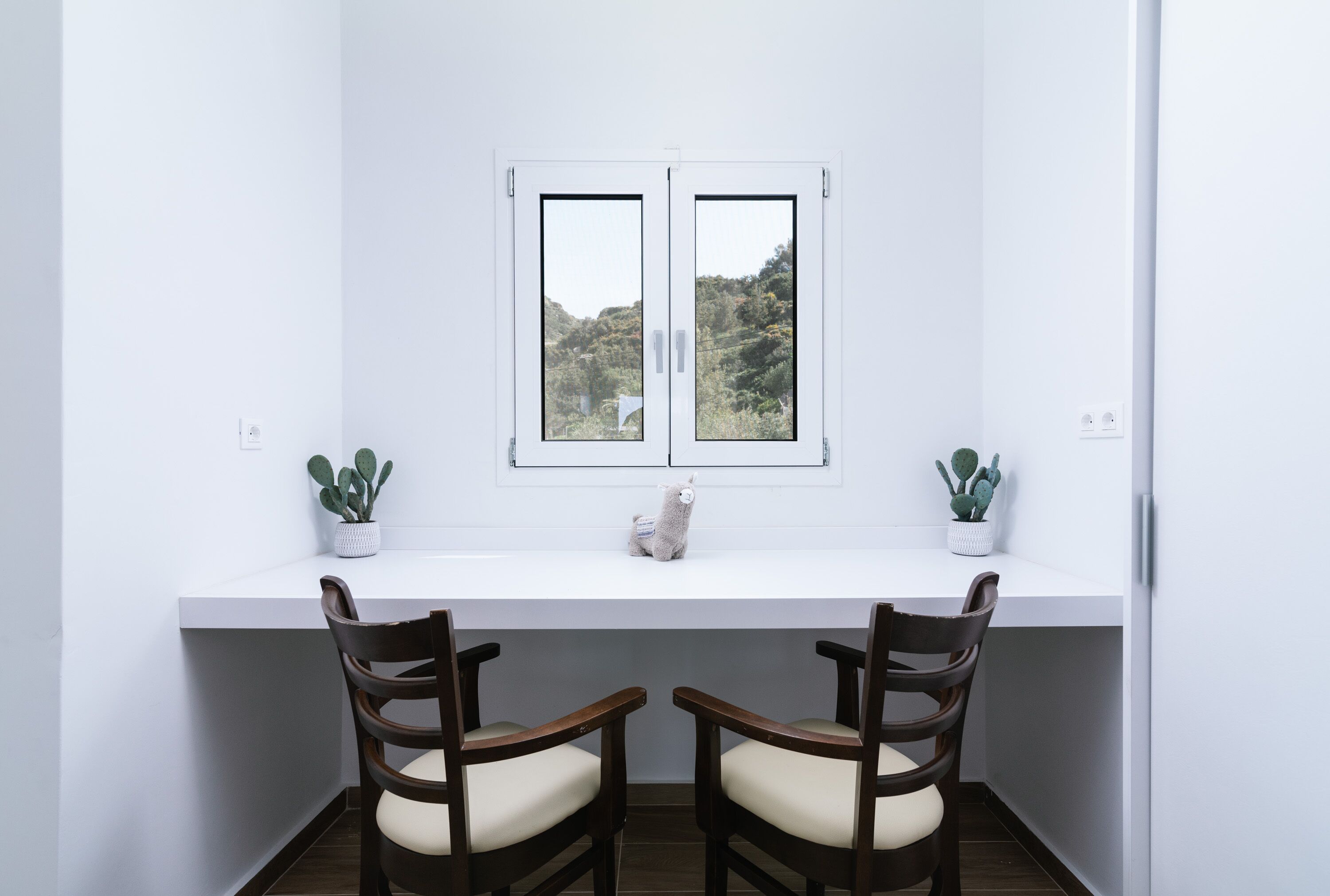 A white desk with two chairs, a window with a view of hills, and two potted cacti.