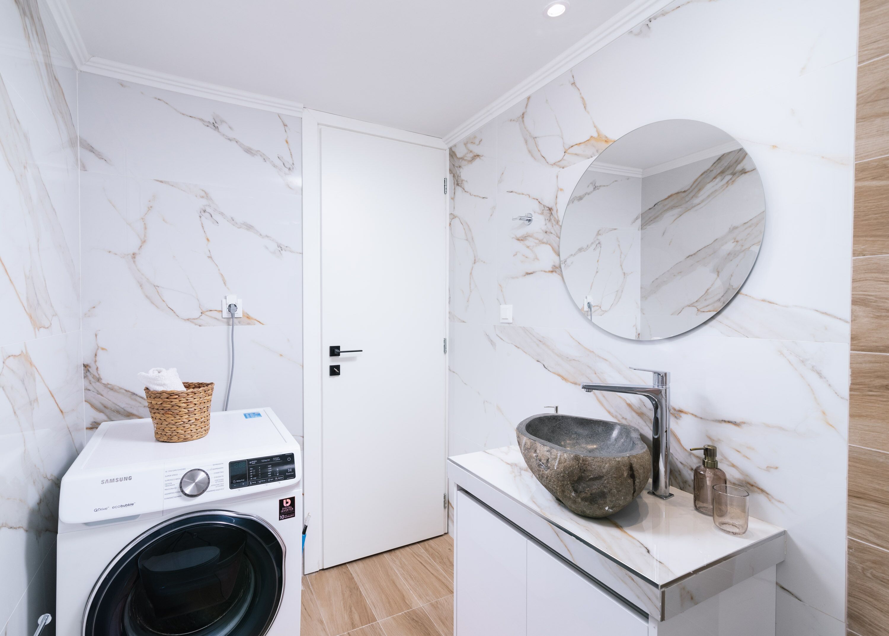 A modern bathroom with a marble wall, a washing machine, a sink with a stone basin, and a round mirror.
