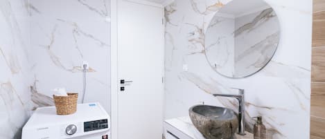 A modern bathroom with a marble wall, a washing machine, a sink with a stone basin, and a round mirror.