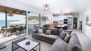 A modern living room with a large gray sofa, a glass dining table, and a view of the sea. - Beach front apartment with swimming pool ideal for families (Ligaria Beach)