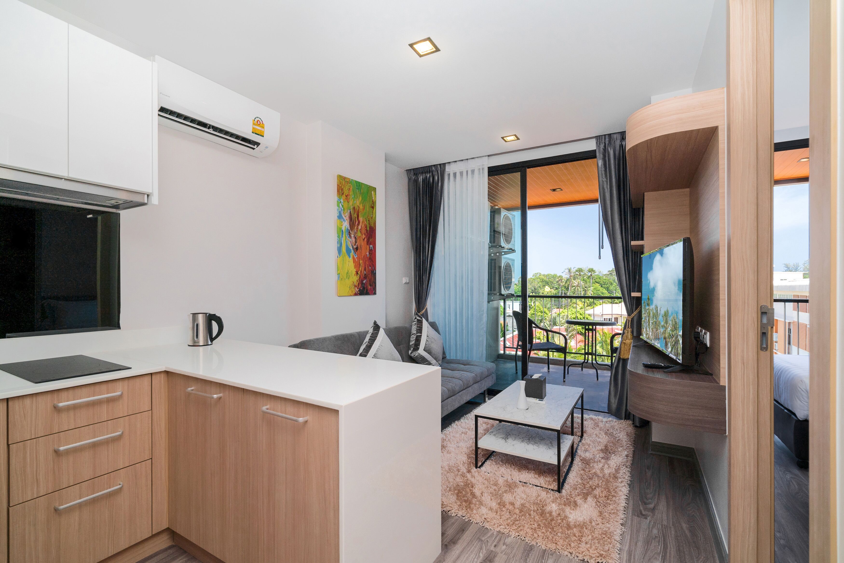 A modern kitchen with white cabinets, a black countertop, and a coffee maker. There is a sofa with a fluffy rug, a TV, and a balcony with a view of the city.