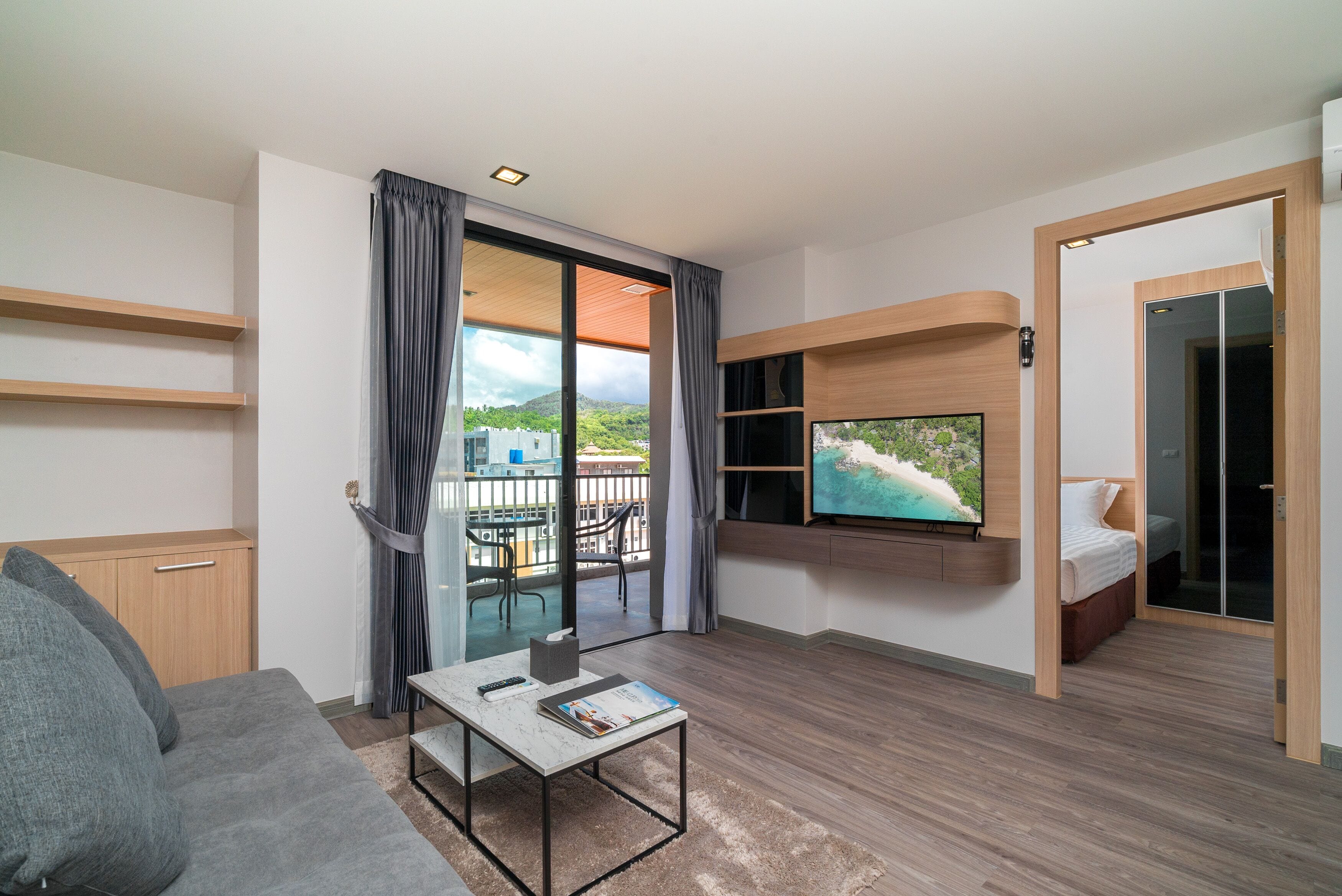 A modern hotel room with a sofa, a small table, a TV mounted on the wall, a balcony with a view of the city, and a sliding door leading to a bedroom.