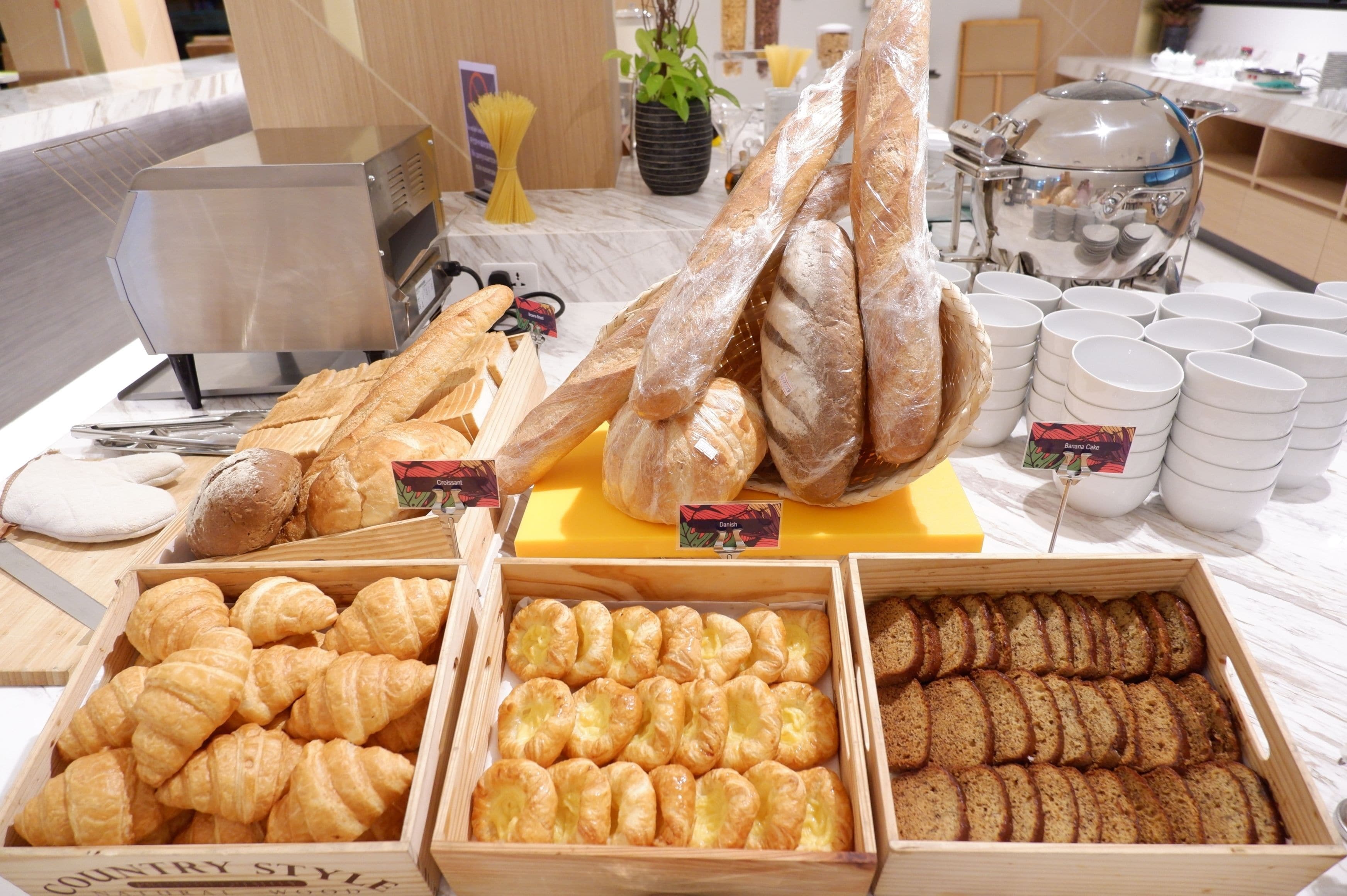 A buffet table with various baked goods, including croissants, bread, and rolls, along with coffee cups and a coffee pot.