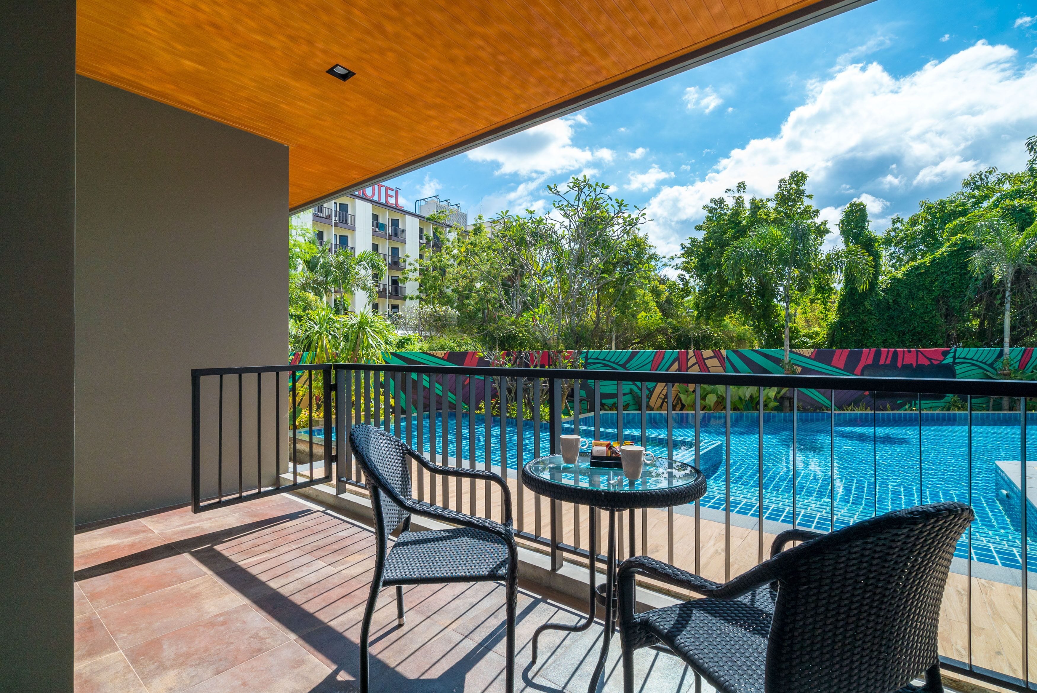 A balcony with wicker chairs and a small table, overlooking a swimming pool and a hotel building.