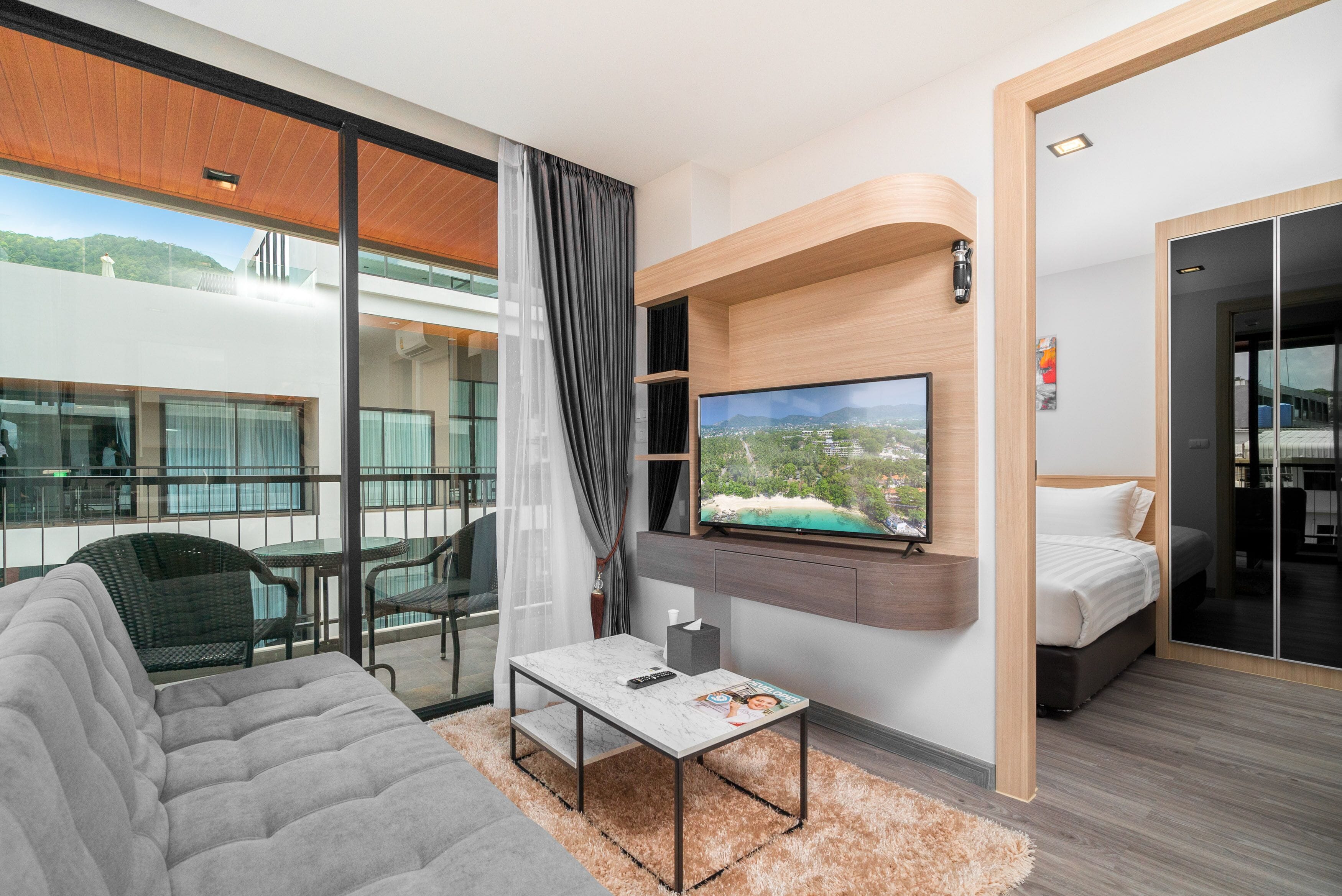 A modern hotel room with a large TV, a sofa, a dining area with chairs, and a balcony with a view of the city.