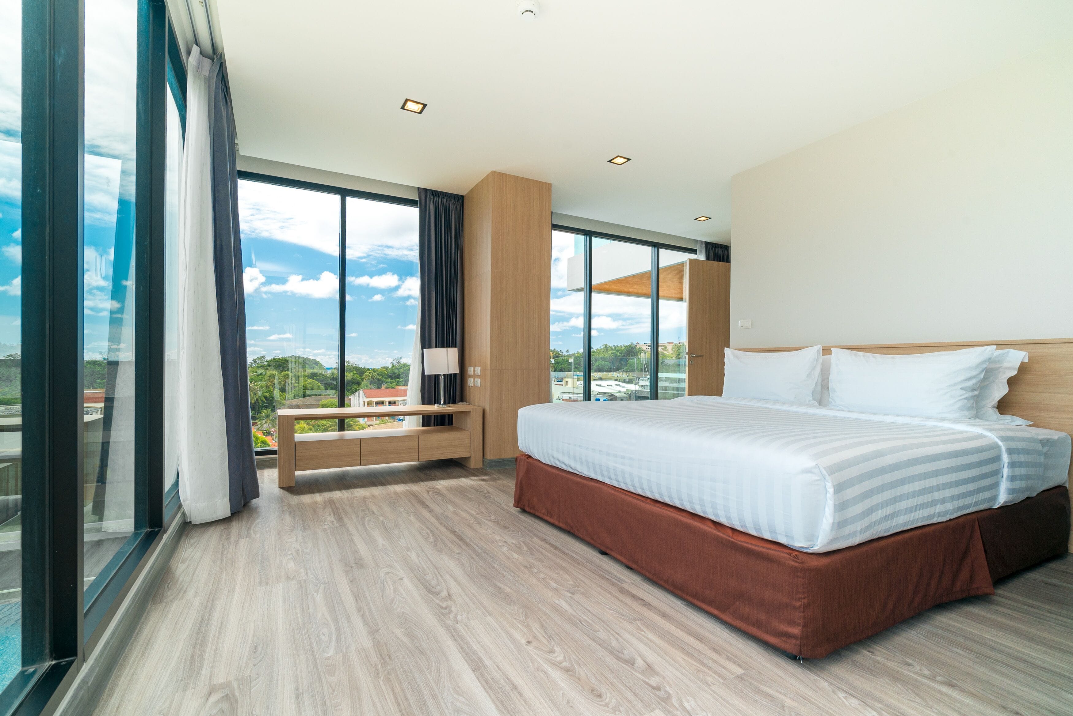 A modern hotel room with a large bed, a nightstand, a bench, and a view of the city.