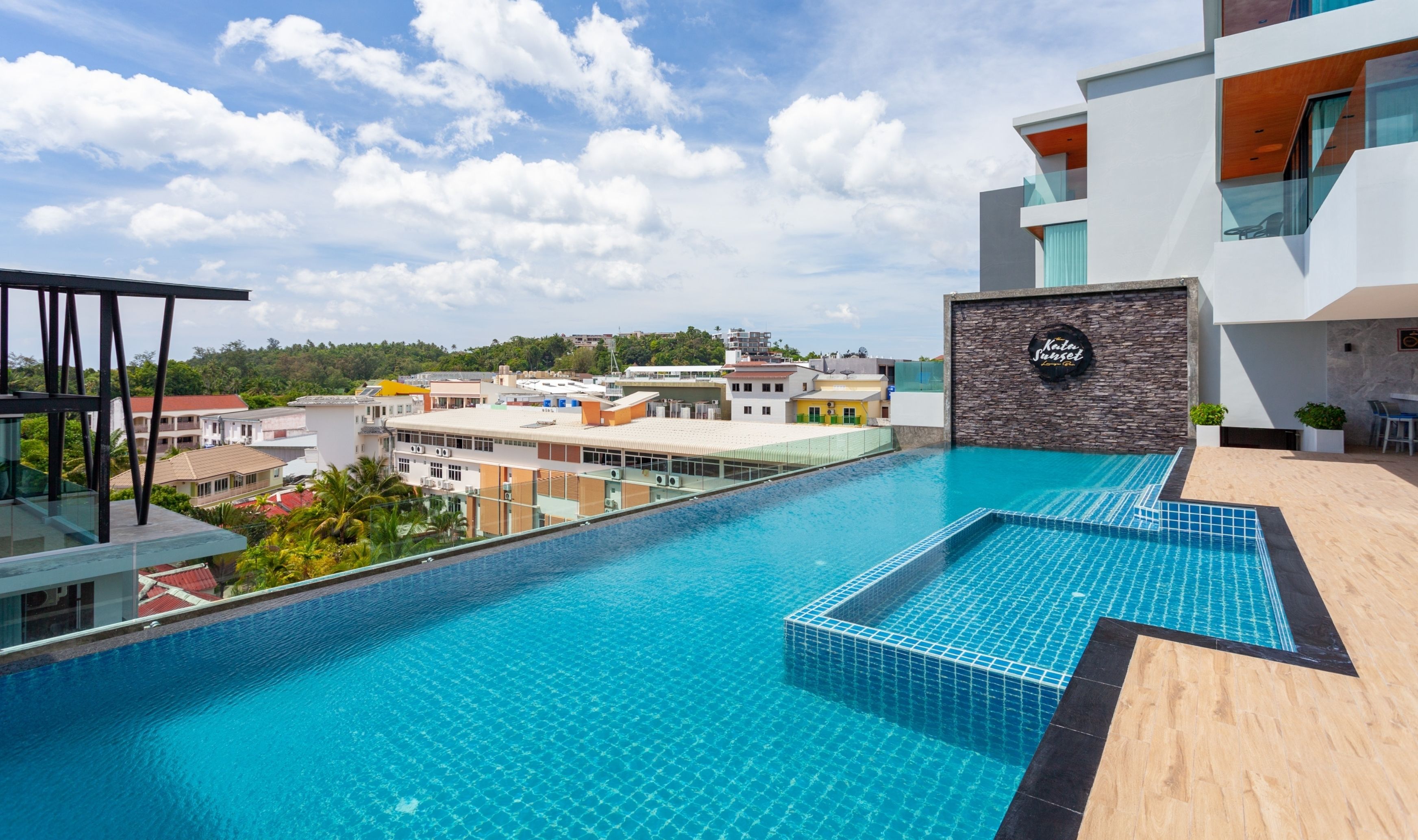 A rooftop infinity pool with a view of the cityscape, a modern building with a stone facade, and a sign that reads "Kata Sunset".
