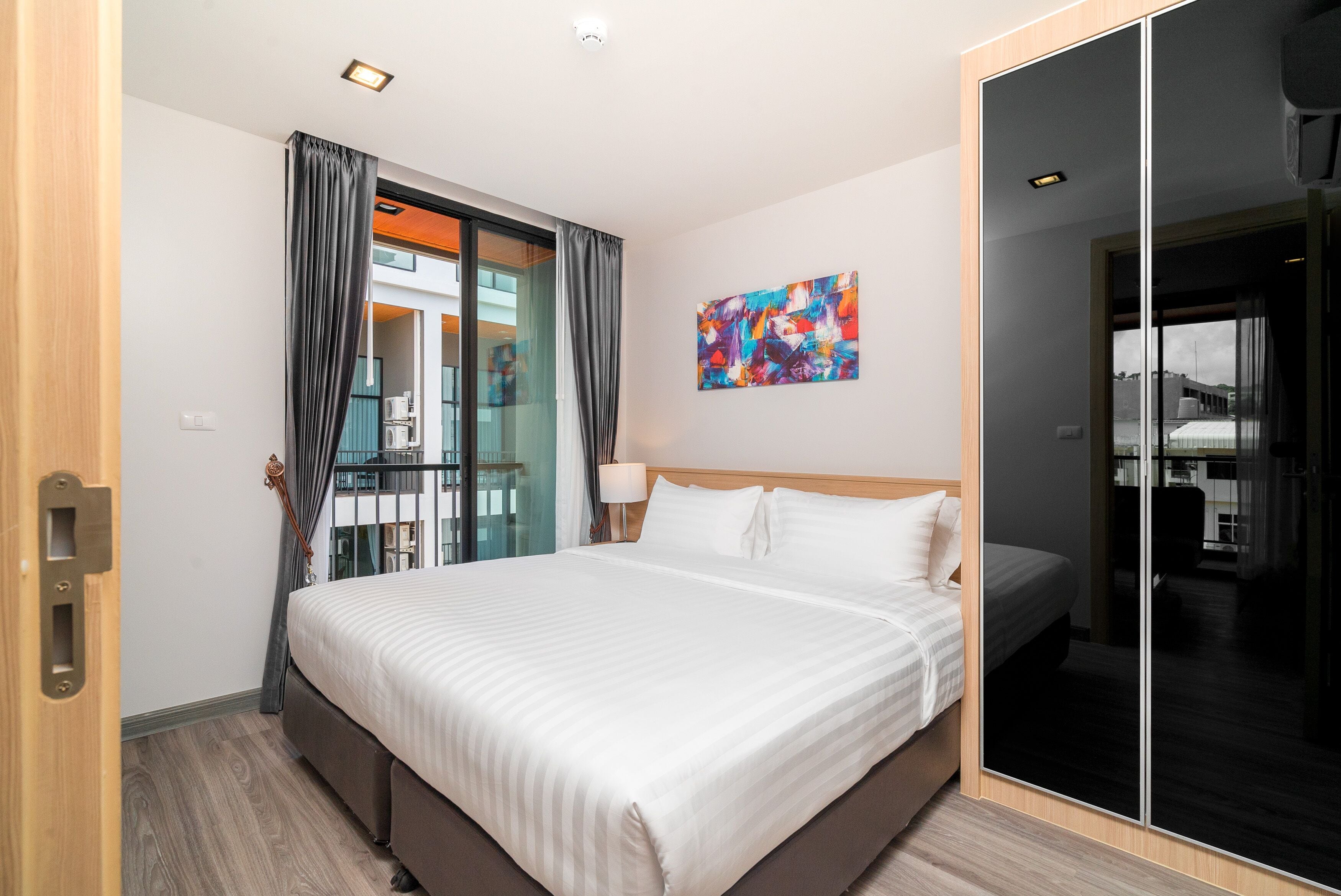 A hotel room with a large bed, a glass door leading to a balcony, a painting on the wall, and a wooden door.