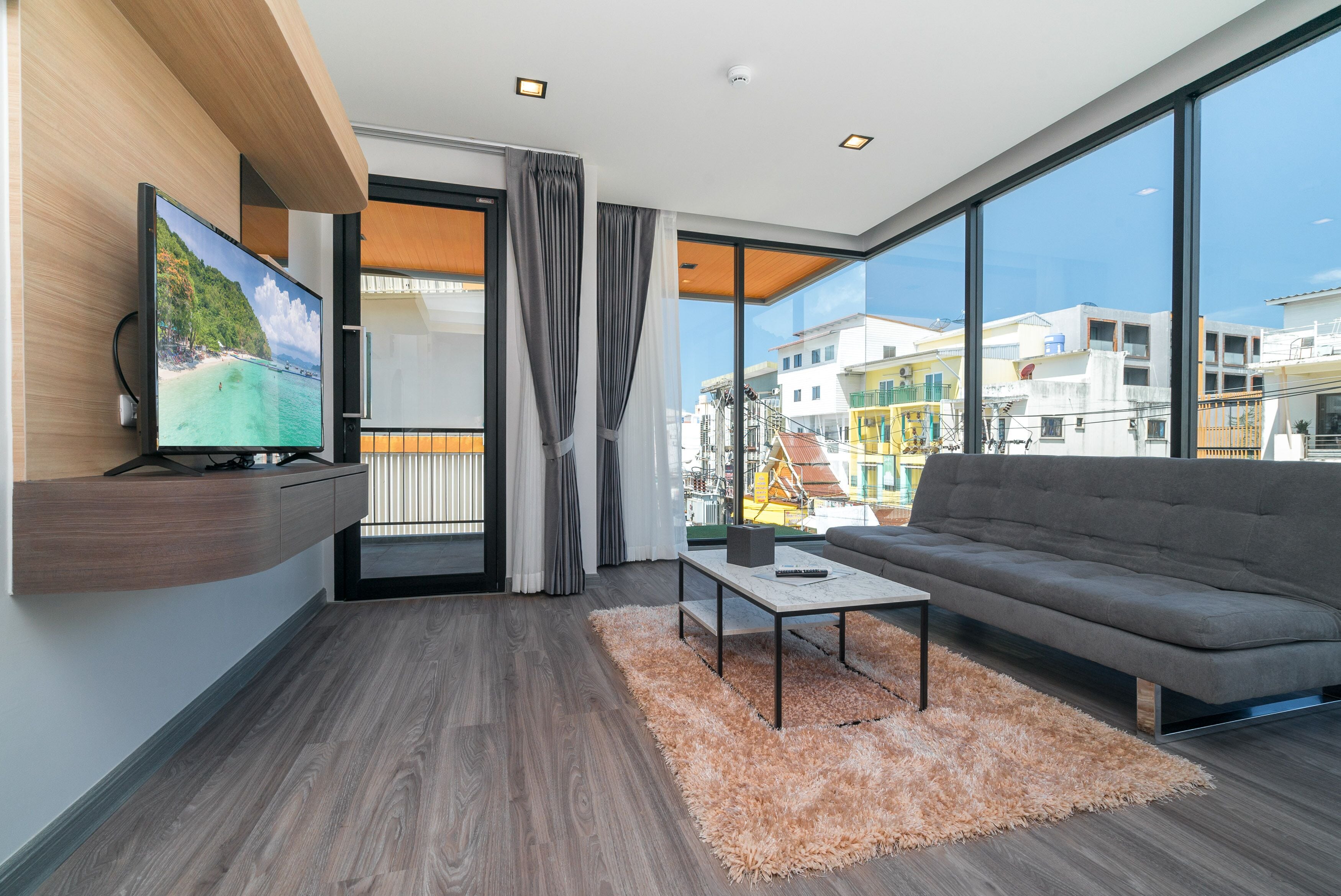 A modern living room with a large TV, grey sofa, and a view of buildings through large windows.