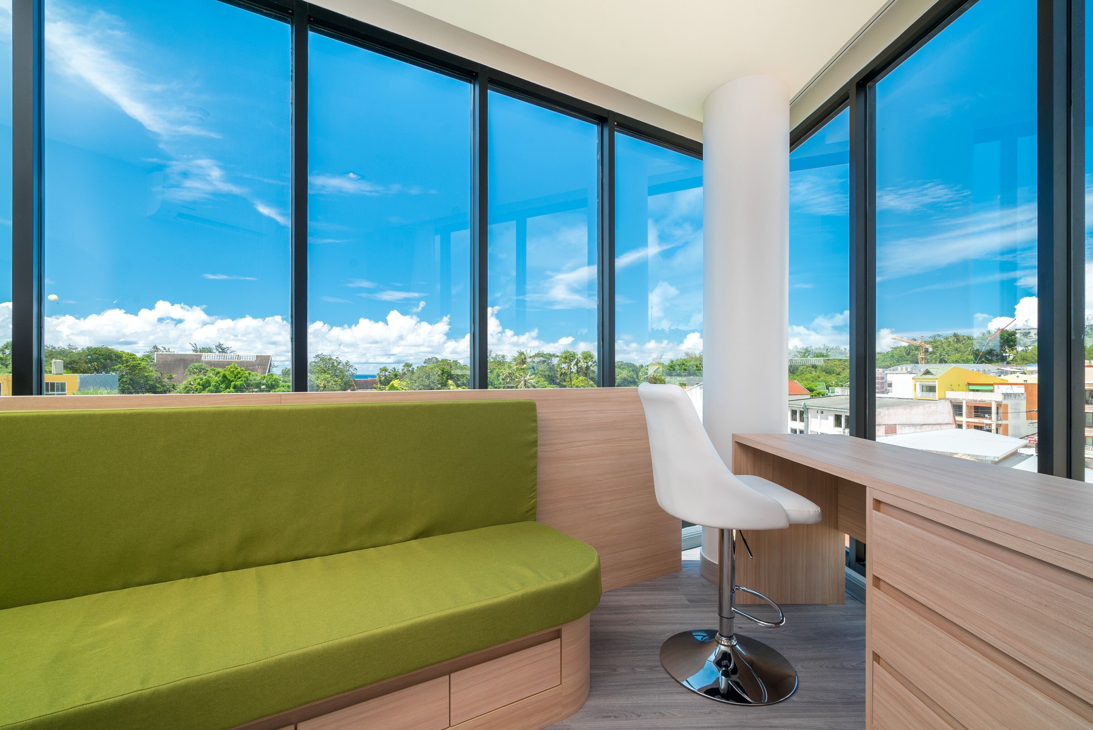 A modern room with a green sofa, a white chair, and a wooden desk. Large windows offer a view of the cityscape.
