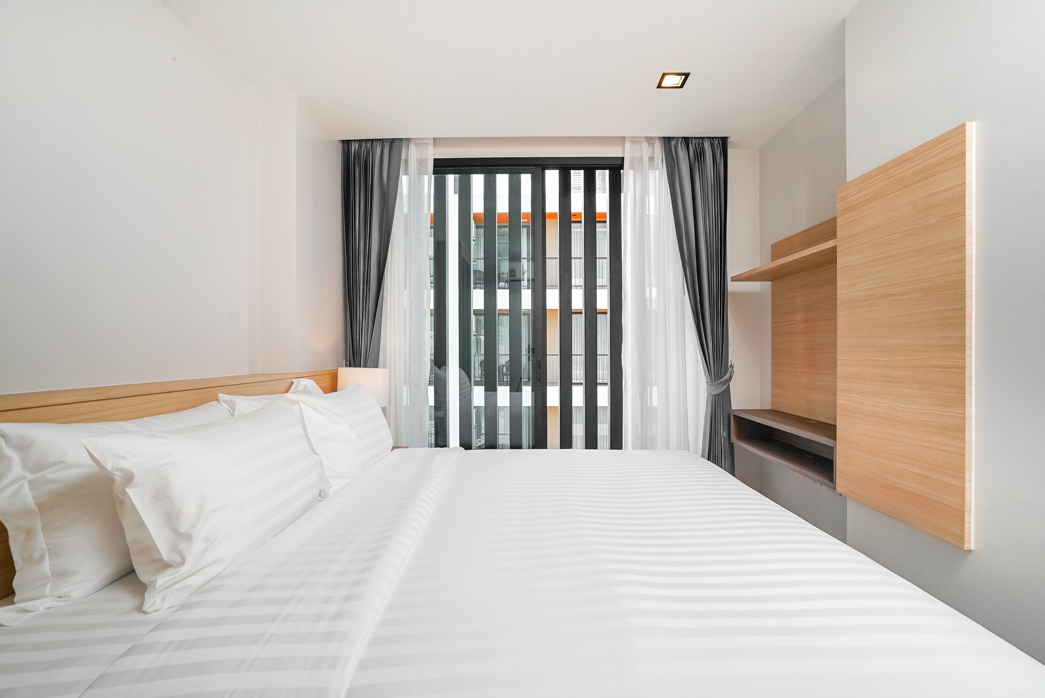 A modern hotel room with a large bed, a wooden headboard, a built-in wooden shelf, and a view of a building through sheer curtains.