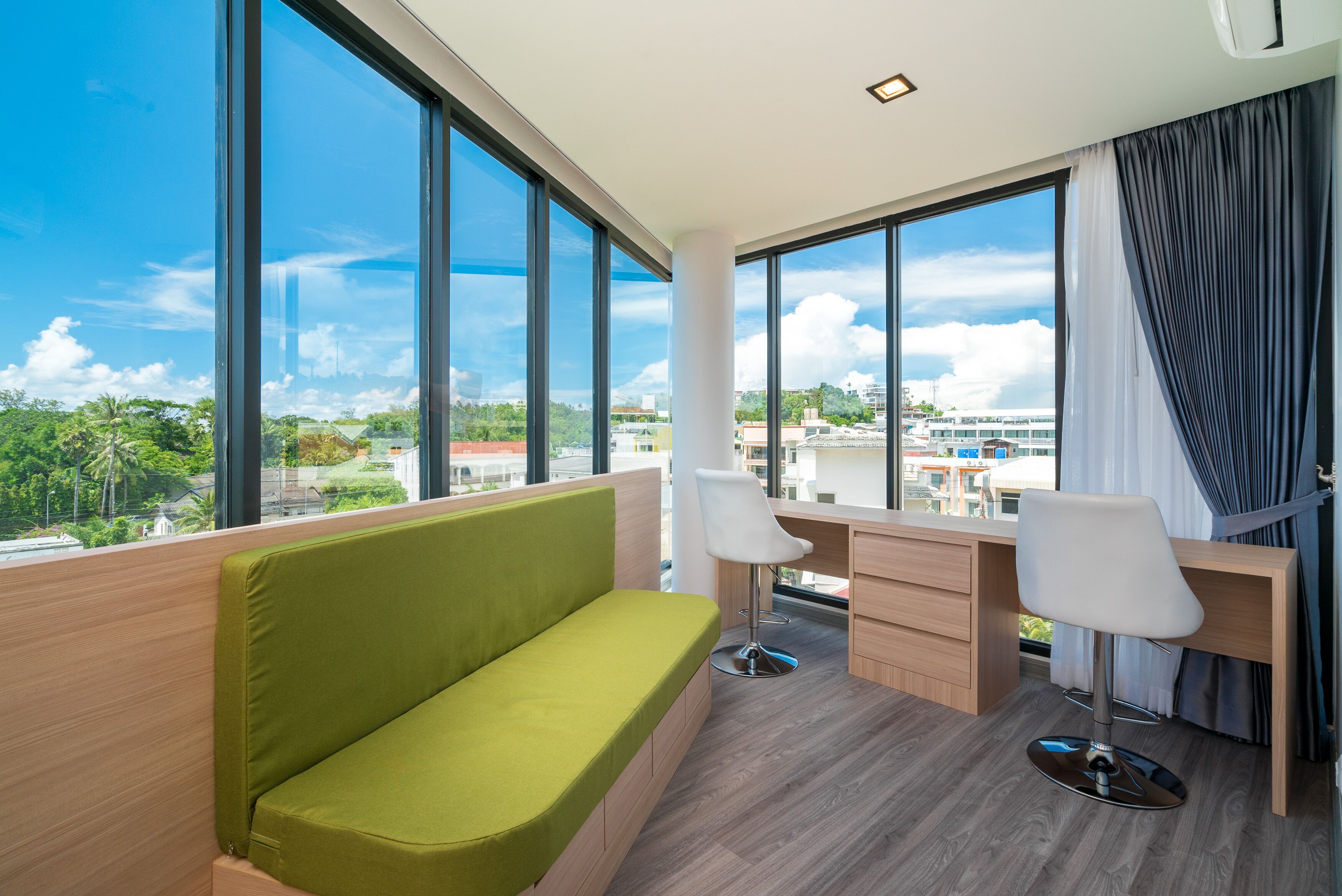 A modern hotel room with a green sofa, two white chairs, a wooden desk, and large windows offering a city view.