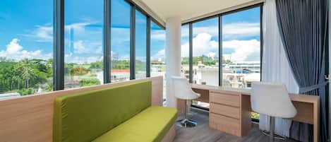 Et moderne hotelværelse med en grøn sofa, to hvide stole, et træskrivebord og store vinduer med udsigt over byen.