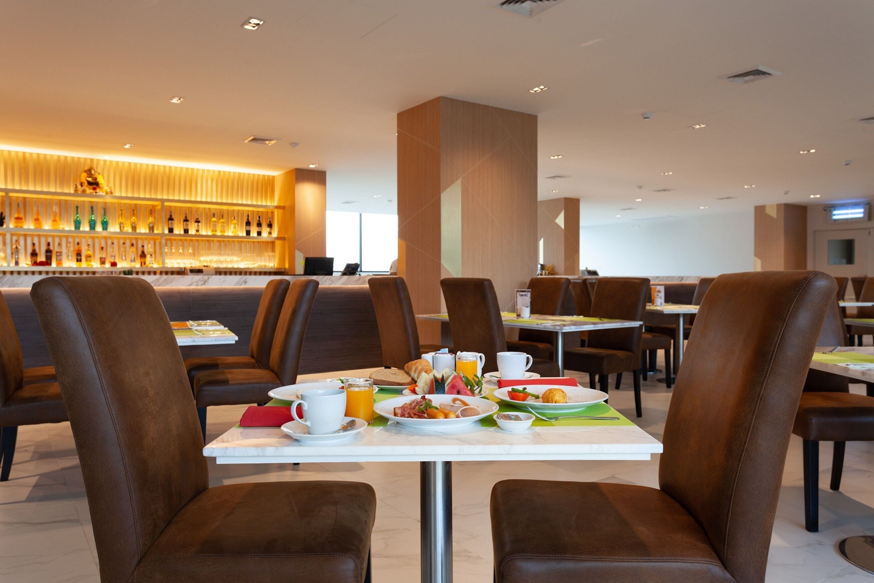 Un espace de restauration d’hôtel avec des fauteuils en cuir marron, un buffet pour le petit-déjeuner et un bar avec des bouteilles exposées.