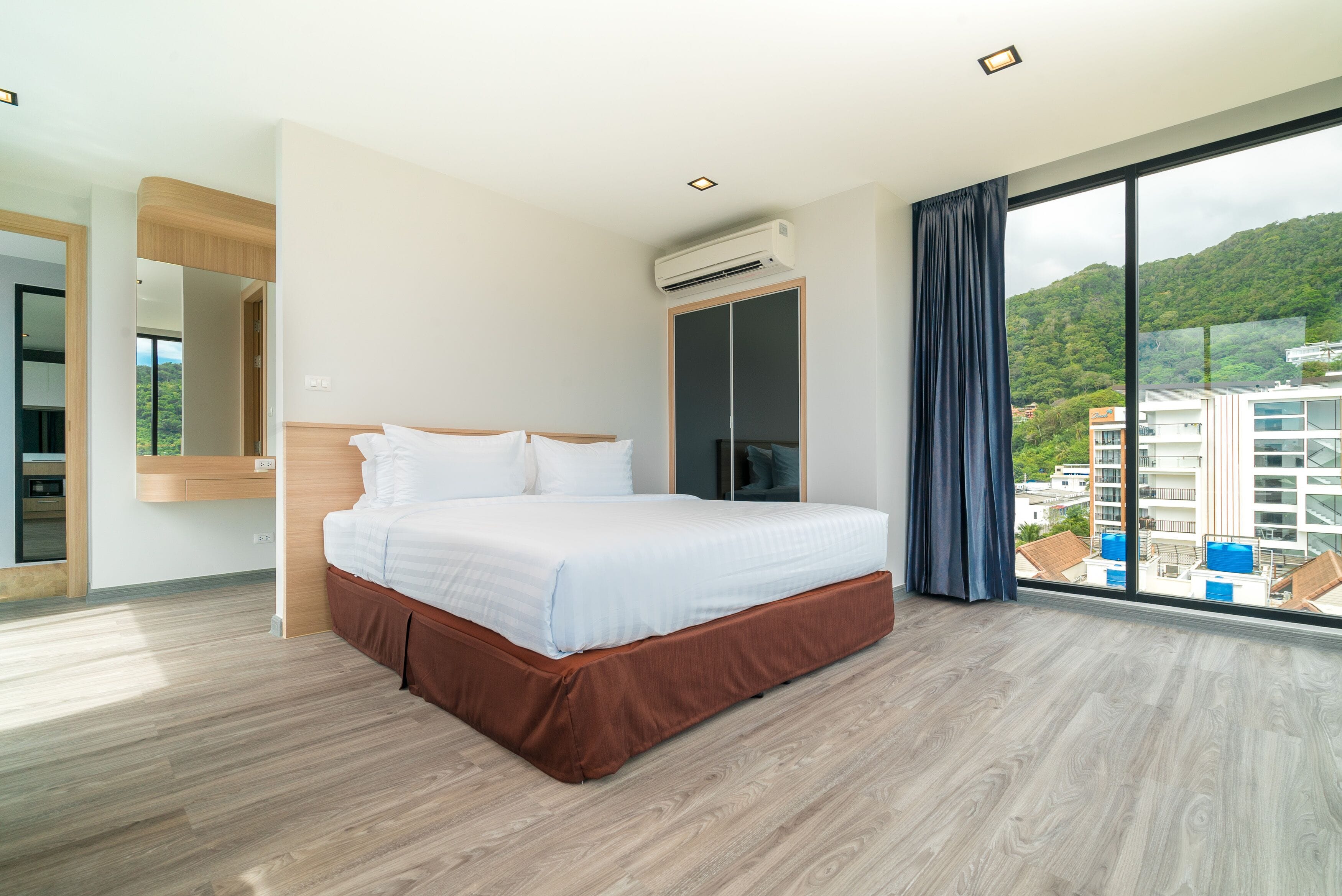 A modern hotel room with a large bed, a wardrobe, an air conditioning unit, and a large window offering a view of buildings and greenery.