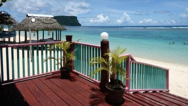 Una terraza de madera con barandilla que da a una playa, una estructura con techo de paja y vista al mar y una isla distante.