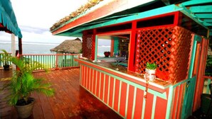 Una terraza de madera con techo de paja, barandilla roja y verde, y vista al océano.