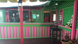 Un espacio al aire libre techado, con piso de madera, barandales verdes y rosados, y una silla.