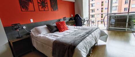 A hotel room with a bed, a desk, and a chair. There is a window with a view of buildings outside.