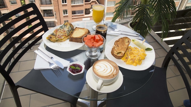 Une table dressée sur un balcon, avec vue sur un immeuble en briques, où sont disposés des éléments pour le petit-déjeuner, notamment des toasts, des œufs brouillés et une tasse de café.