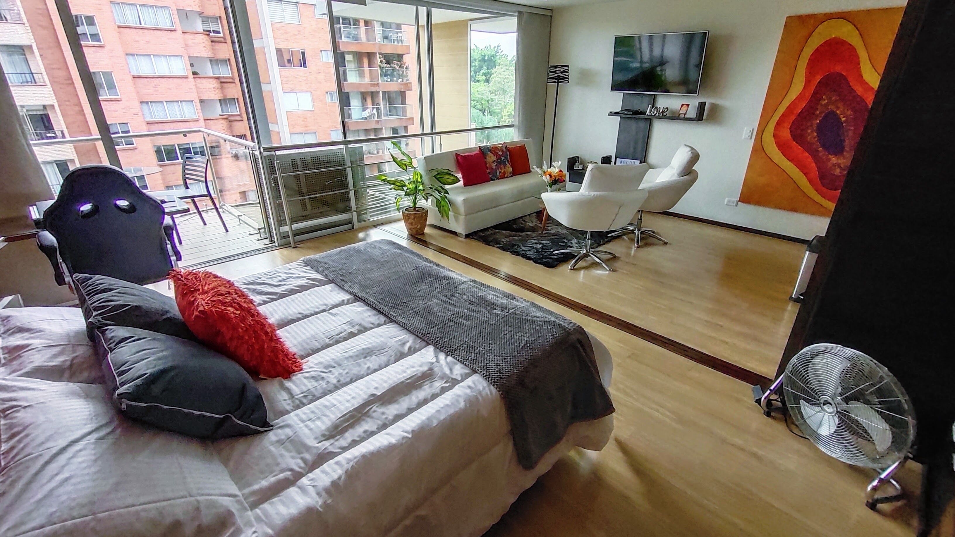 A modern hotel room with a large bed, a sofa, a chair, a TV, and a balcony with a view of the city.