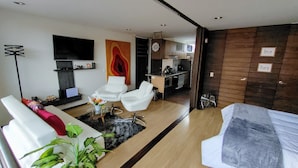 Un salon moderne avec un canapé, deux fauteuils blancs, une table basse et un téléviseur. On aperçoit une cuisine à l’arrière-plan.